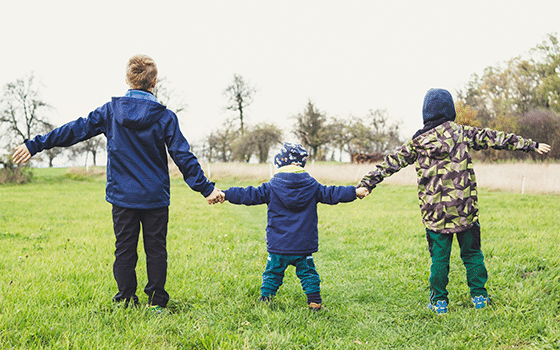 three children holding hands while facing away from the camera in a park