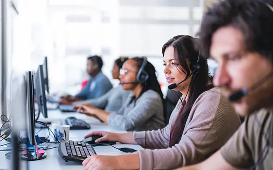 Young businesswoman wearing headset while using computer. Row of operators are sitting at desk. They are working in call centre.